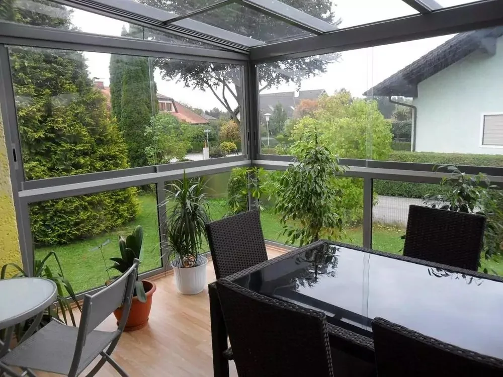 Terrassendach mit Glaswänden, darunter ein Esstisch mit Rattanstühlen auf Holzboden